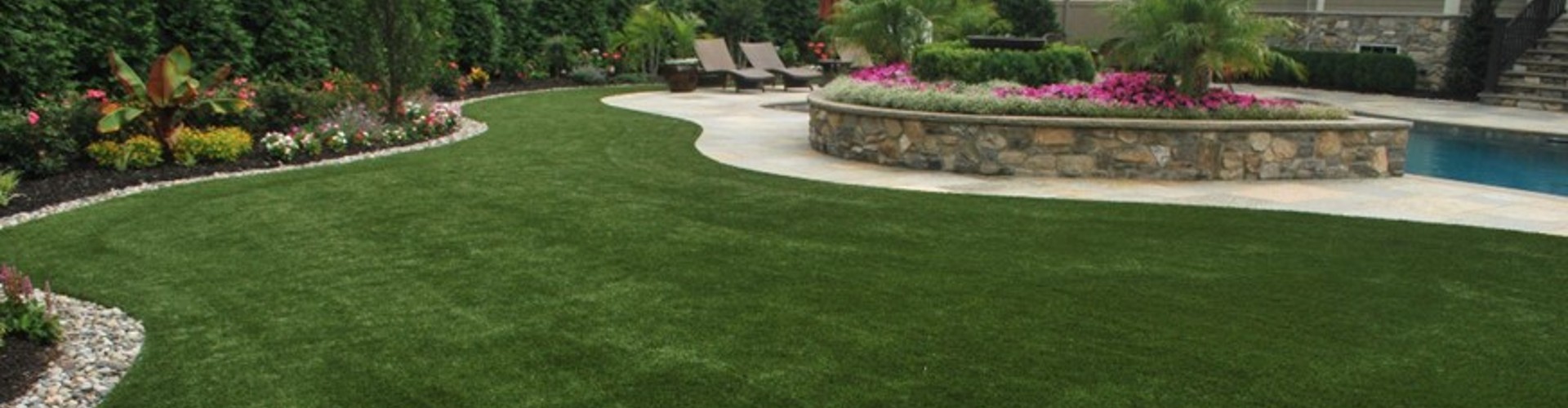 Southwest Greens lawn turf grass flowing around a stone raised garden planter, pool edge, and colorful tropical flower beds with lounge chairs.