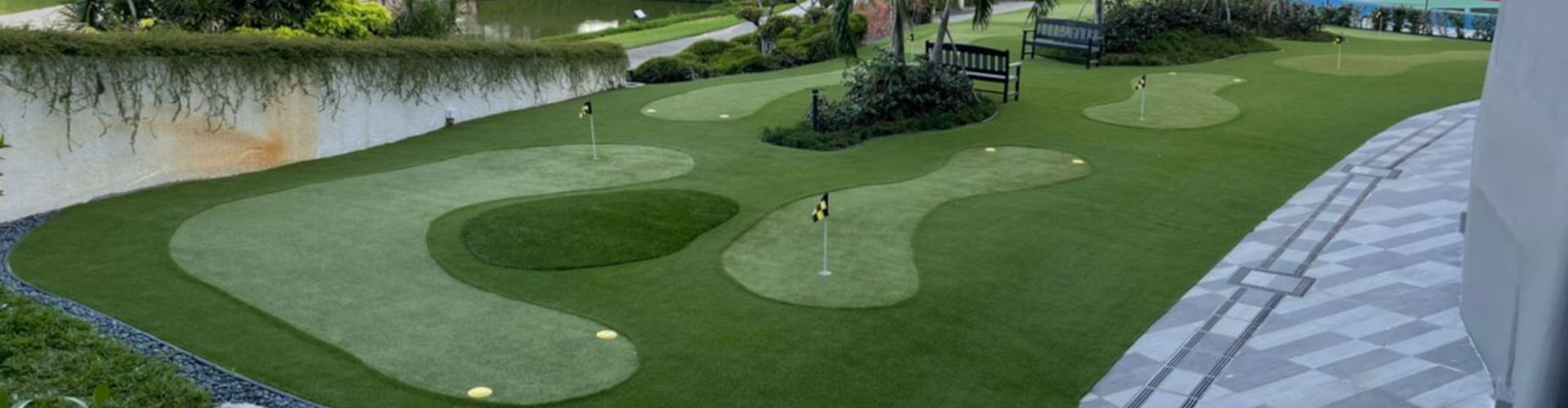 Multi-hole putting green turf installation at a luxury golf club with varied cup targets, yellow flag pins, lush tropical landscaping, and a decorative tile walkway border