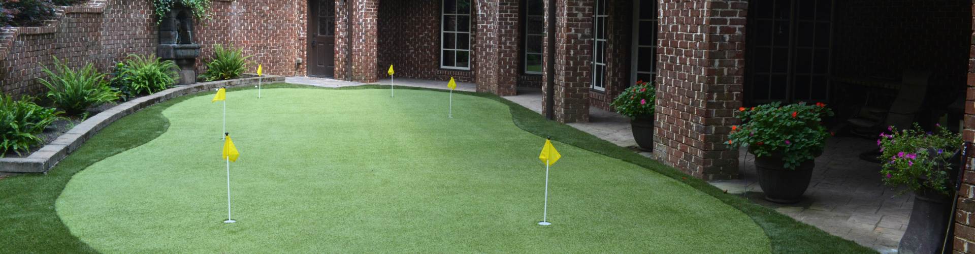 Custom backyard putting green with five yellow flag pins installed on a brick home terrace with surrounding garden landscaping